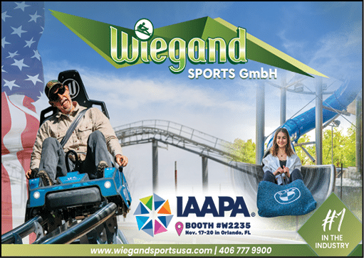 Two people are enjoying a ride on a blue amusement park attraction with a roller coaster and water slide in the background, alongside promotional text for Wiegand Sports GmbH and IAAPA event details. Description generated by AI