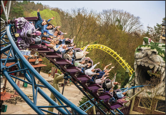 People with raised arms are riding a roller coaster descending a steep track near a large rock structure with a carved face. Description generated by AI