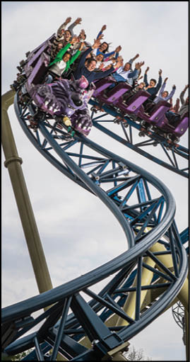 People with raised hands are riding a purple roller coaster car shaped like a creature on a twisting blue track against a cloudy sky. Description generated by AI