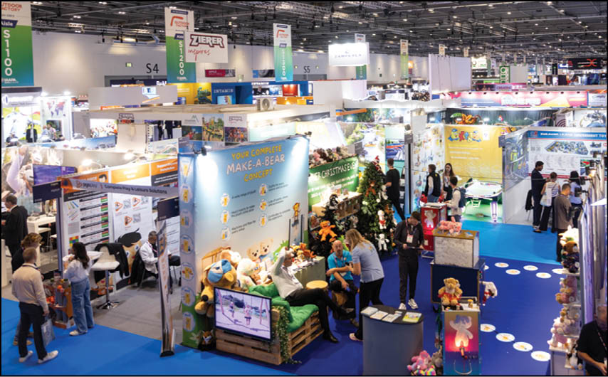 A busy indoor convention or trade show features multiple booths with colorful displays, people walking and interacting, and a prominent booth showcasing stuffed bears and a sign about a Make-A-Bear concept. Description generated by AI