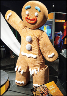 A large gingerbread man costume with blue eyebrows, white eyes, red mouth, and white icing decorations stands indoors near informational displays. Description generated by AI