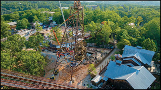 A tall metal structure resembling a mining or industrial tower stands in a wooded area surrounded by buildings and pathways. Description generated by AI