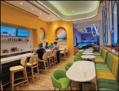 A modern bar and lounge area with people seated at the counter and colorful seating along the wall, featuring bright yellow and green decor and multiple TV screens. Description generated by AI