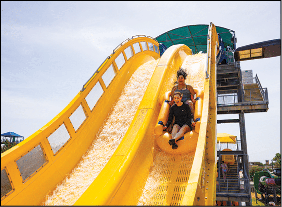 Two people are riding a yellow inflatable raft down a bright yellow water slide at a water park on a sunny day. Description generated by AI