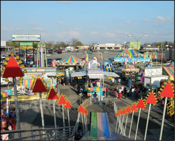 A colorful outdoor carnival scene with tents, rides, ticket booths, and people walking around under a clear sky. Description generated by AI