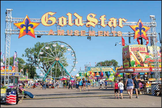 A lively outdoor amusement park scene with a large Gold Star Amusements sign, a Ferris wheel, colorful game booths, and people walking along the pathway under a clear blue sky. Description generated by AI