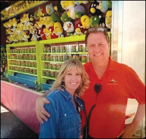 Two adults smiling and posing together in front of a carnival game booth filled with stuffed animal prizes. Description generated by AI