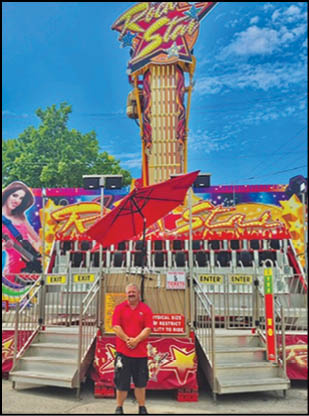 A person wearing a red shirt and black shorts stands in front of a colorful amusement ride with a red umbrella overhead and signs indicating ticket entry and exit. Description generated by AI