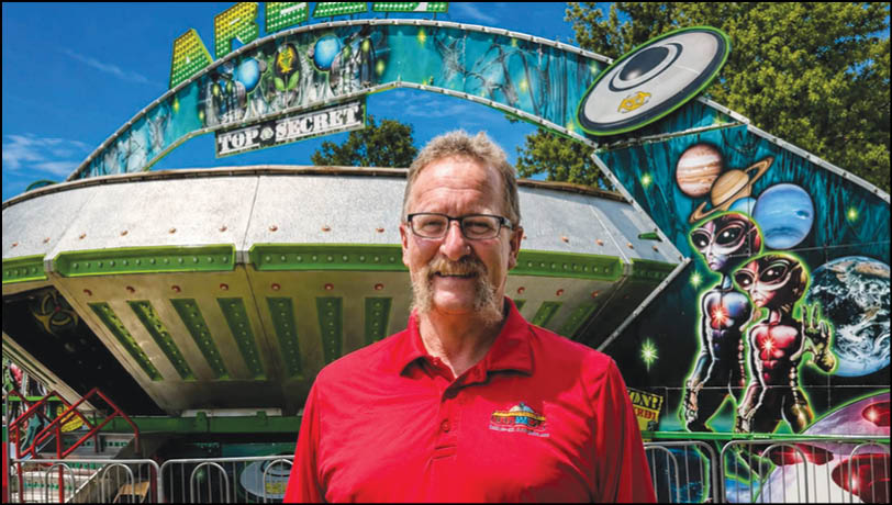 A person wearing glasses and a red shirt stands in front of a colorful amusement ride decorated with alien and space-themed artwork. Description generated by AI