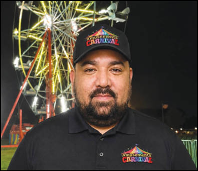 A person wearing a black polo shirt and black cap with colorful “Amusements Carnival" logos stands in front of a brightly lit Ferris wheel at night. Description generated by AI