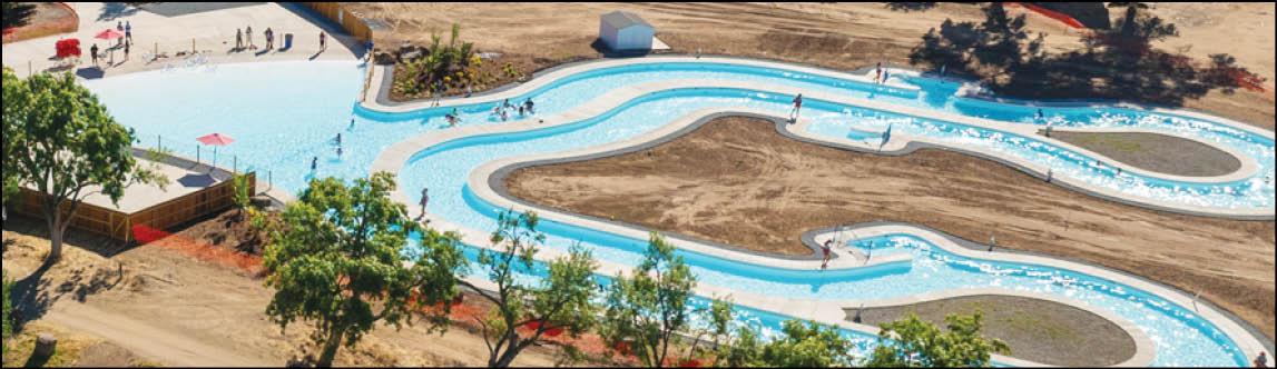 A winding outdoor water slide with several people sliding and walking around in a sunny, open area surrounded by trees and dry ground. Description generated by AI
