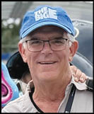 A senior person wearing glasses and a blue cap with the words “THE BIG E" smiles while standing outdoors with a hand resting on their shoulder. Description generated by AI