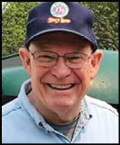A smiling senior wearing a cap and glasses stands next to a small red train car labeled “C.P. Hunting" in an outdoor park setting. Description generated by AI