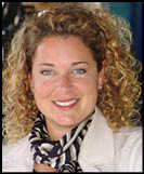 A person with curly hair is smiling and wearing a light-colored jacket and a black and white striped scarf. Description generated by AI