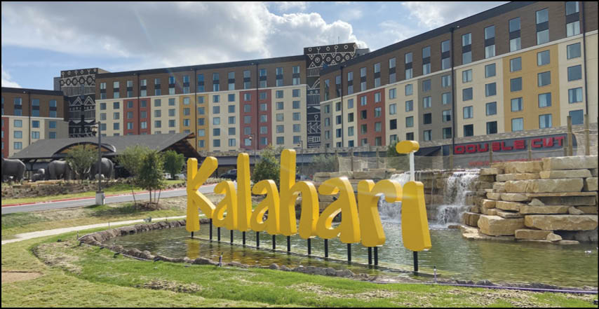 A large yellow sign spelling “Kalahari" stands in front of a pond with a waterfall, with a multi-story building and a sign reading "Double Cut" in the background. Description generated by AI
