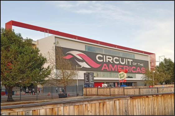 A large building with a prominent sign reading “Circuit of the Americas" is surrounded by trees and a wooden fence under a partly cloudy sky. Description generated by AI
