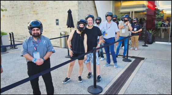 A group of people wearing helmets and casual clothing are standing in a line outdoors behind a black rope barrier near a building entrance. Description generated by AI
