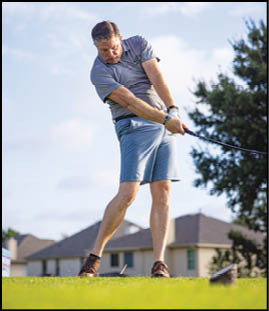 A person wearing a gray shirt and blue shorts is swinging a golf club on a grassy area with houses and a tree in the background. Description generated by AI