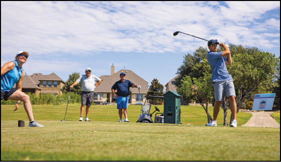 Four people in casual sportswear are playing golf on a sunny day in a residential neighborhood with houses and trees in the background. Description generated by AI