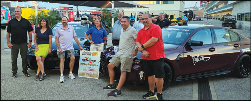 A group of adults stands and sits casually around two parked cars under a canopy in an outdoor setting with buildings and other vehicles in the background. Description generated by AI