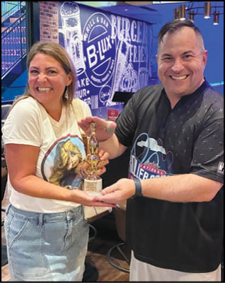 Two adults smiling and holding a trophy together inside a casual dining or bar setting with a mural on the wall behind them. Description generated by AI