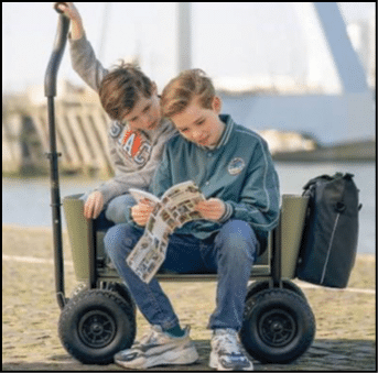 Two children sit on a small four-wheeled cart by the water, with one child reading a newspaper and the other holding onto a handle behind them. Description generated by AI