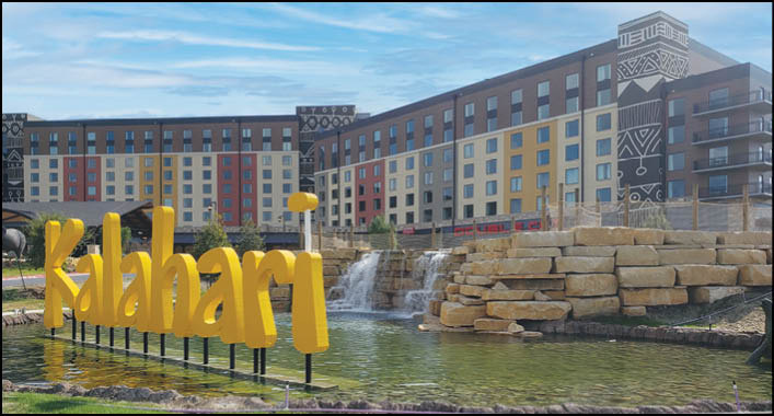 A large modern building with colorful panels and decorative patterns is situated behind a pond with a waterfall and a yellow sign spelling “Kalahari." Description generated by AI