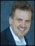 A person with tousled hair wearing a dark blazer and a light checkered shirt is smiling against a plain dark background. Description generated by AI