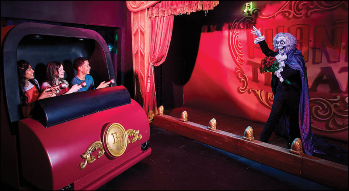 Several children seated in a red ride vehicle watch and react to a costumed performer dressed in a dark cape and white wig who is gesturing dramatically on a stage with red curtains and lighting. Description generated by AI