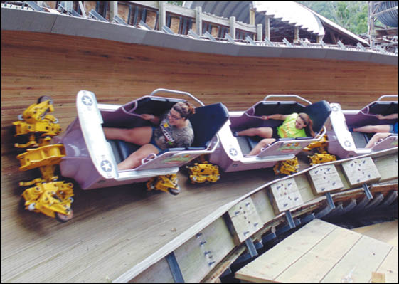 People wearing casual clothing are seated in a tilted roller coaster car on a wooden track, appearing to be in motion. Description generated by AI