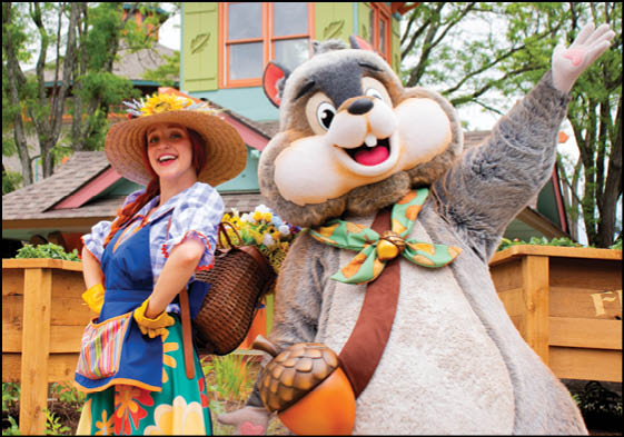 A person wearing a colorful floral dress, apron, and wide-brimmed hat stands smiling next to a large chipmunk mascot holding an acorn and waving in an outdoor setting with a wooden structure and trees in the background. Description generated by AI