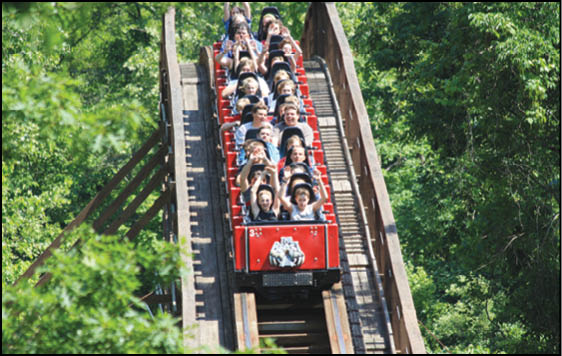 A group of people are seated in a red roller coaster car descending a wooden track surrounded by green trees. Description generated by AI