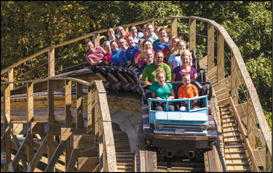 A group of people wearing colorful shirts are riding a wooden roller coaster through a forested area, appearing excited and joyful. Description generated by AI