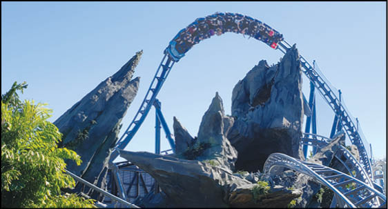 A roller coaster train is descending a steep curved track surrounded by rocky structures and greenery under a clear blue sky. Description generated by AI