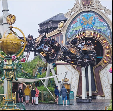 People are riding a spinning amusement park ride shaped like a clock keyhole with ornate decorations and stained glass, while others stand nearby under an overcast sky. Description generated by AI