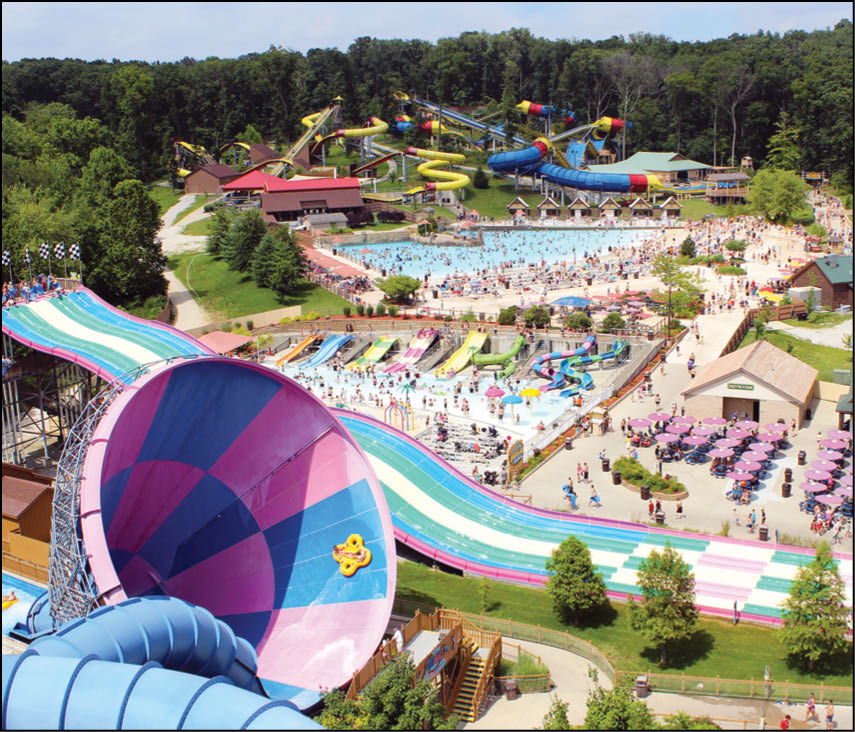 A crowded water park with multiple colorful water slides, pools filled with people, and surrounding trees under a clear sky. Description generated by AI