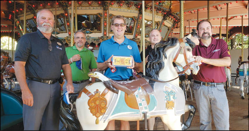 Five adults stand around a decorative carousel horse inside a carousel structure, with one person holding a small rectangular object. Description generated by AI