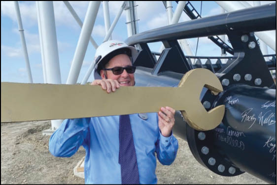 A person wearing a blue shirt, tie, safety helmet, and sunglasses is holding a large golden wrench prop near industrial equipment outdoors. Description generated by AI