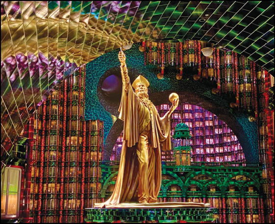A golden statue of a robed figure holding a sphere and raising one hand stands in front of a colorful, intricate background with geometric and architectural patterns. Description generated by AI
