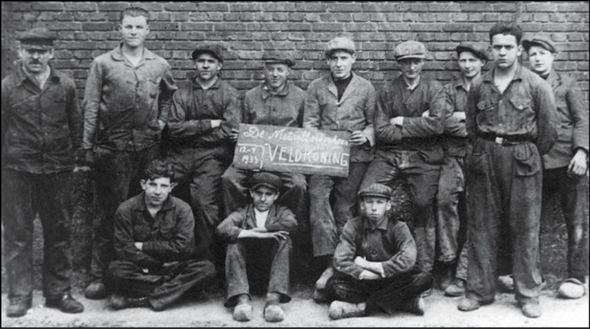 A group of people dressed in work clothes pose in front of a brick wall with one person holding a sign with text. Description generated by AI