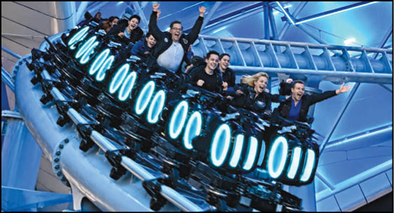 A group of people wearing dark clothing are excitedly riding a roller coaster inside a brightly lit indoor amusement park. Description generated by AI