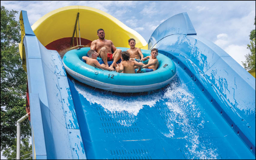 A group of people is riding together on a blue inflatable raft down a large blue water slide with a yellow tunnel at the top. Description generated by AI