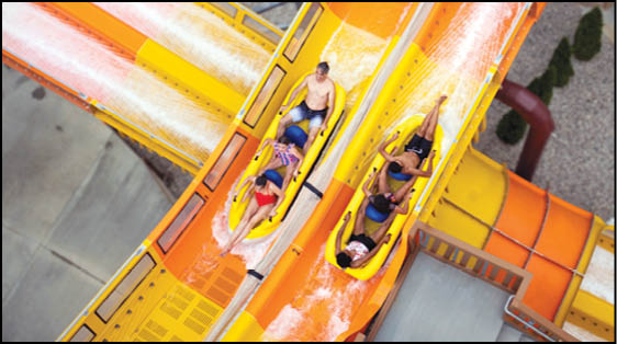 Two people are riding together on each of two yellow water slides at a water park, surrounded by orange and yellow slide structures. Description generated by AI