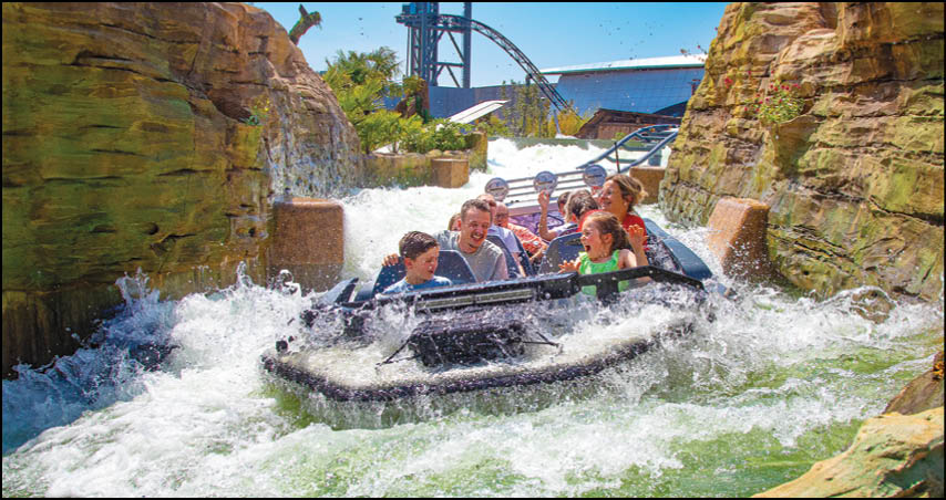 A group of people wearing casual clothes are riding a water raft through a rocky, narrow water ride with splashing waves and a roller coaster visible in the background. Description generated by AI