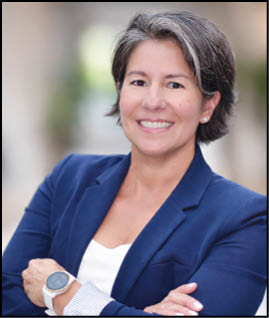 A person with short hair wearing a blue blazer and white top is smiling with arms crossed in a softly blurred outdoor setting. Description generated by AI