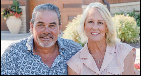 Two adults with light-colored hair are sitting closely together outdoors, smiling, with plants and a building in the background. Description generated by AI