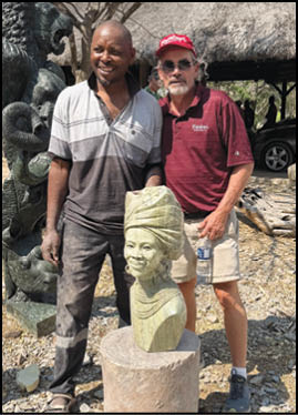 Two adults stand outdoors next to a carved stone bust of a person wearing a traditional headwrap and necklace, with a thatched roof structure and sculptures in the background. Description generated by AI