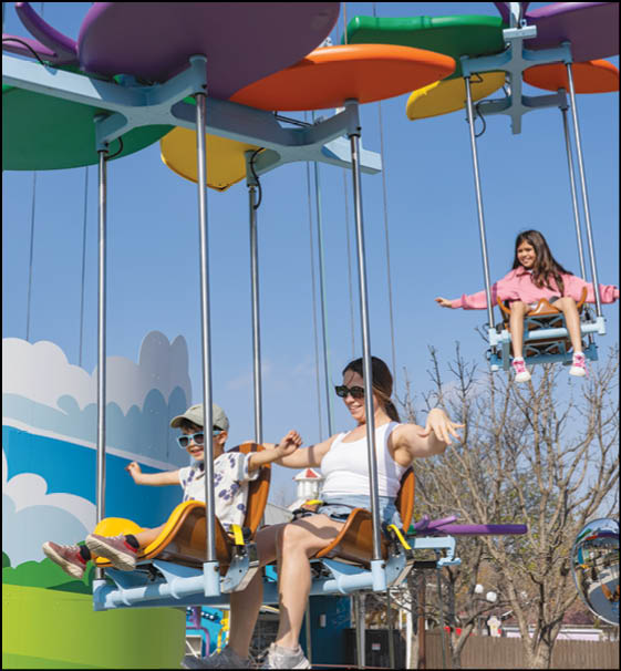 Three people are enjoying a colorful swing ride outdoors on a clear day with blue skies and bare trees in the background. Description generated by AI