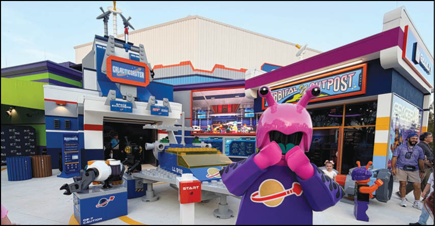 A person in a purple alien costume with pink headgear stands in front of a colorful themed attraction area with signs, interactive displays, and several other people walking nearby. Description generated by AI