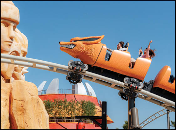 A roller coaster designed like an orange fox with people riding it passes near large stone sculptures of faces against a clear blue sky. Description generated by AI